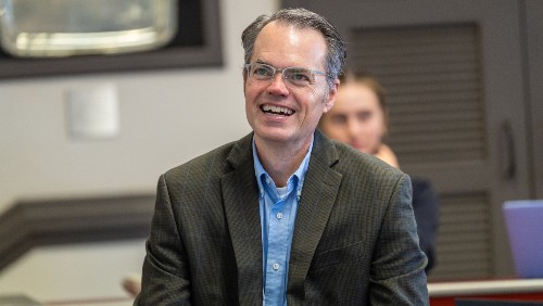 An instructor wearing a blue dress shirt and dark blazer grins in a classroom.